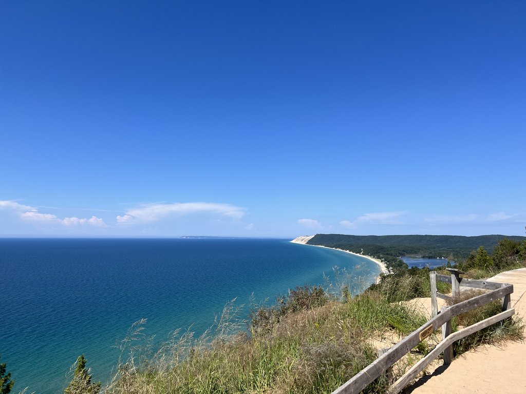 Lake Michigan and a curving sand bluff seen from a dune-top overlook at Sleeping Bear Dunes, a split-rail fence in the foreground.